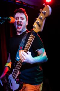 Charlie is in 3/4 profile playing a guitar with his mouth open singing into a mic with an orange covering. He is wearing a black t-shirt and orange slacks.