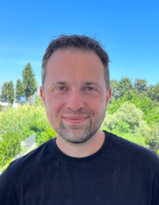 The head and shoulders of Nico appear in front of a background of blue sky and green trees. Nico is wearing a dark blue shirt.